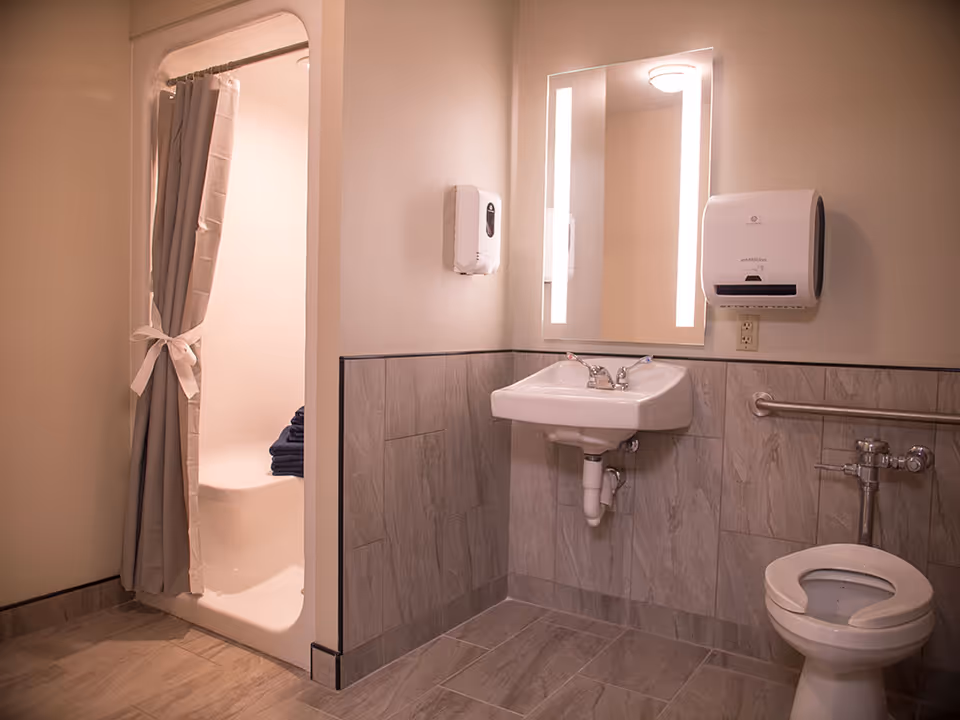 A clean and accessible bathroom featuring a toilet with a safety grab bar, a wall-mounted sink with a lit mirror above it, a soap dispenser, a paper towel dispenser, and a shower area with a gray curtain tied back and folded dark towels inside.