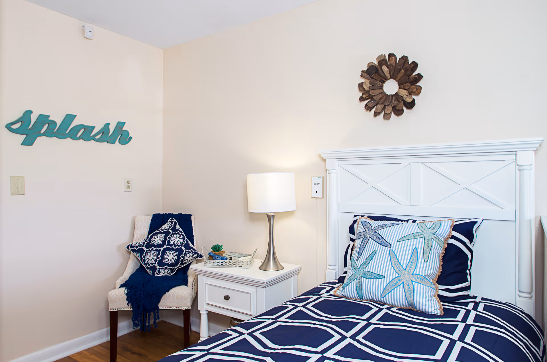 A cozy bedroom corner featuring a white wooden bed with navy blue and white bedding adorned with starfish patterns. Next to the bed is a white nightstand with a modern lamp and a small decorative tray. A beige upholstered chair with a navy blue throw and patterned pillow sits against the wall, which has a teal decorative sign that reads 'splash'. A rustic wooden wall decoration hangs above the bed.
