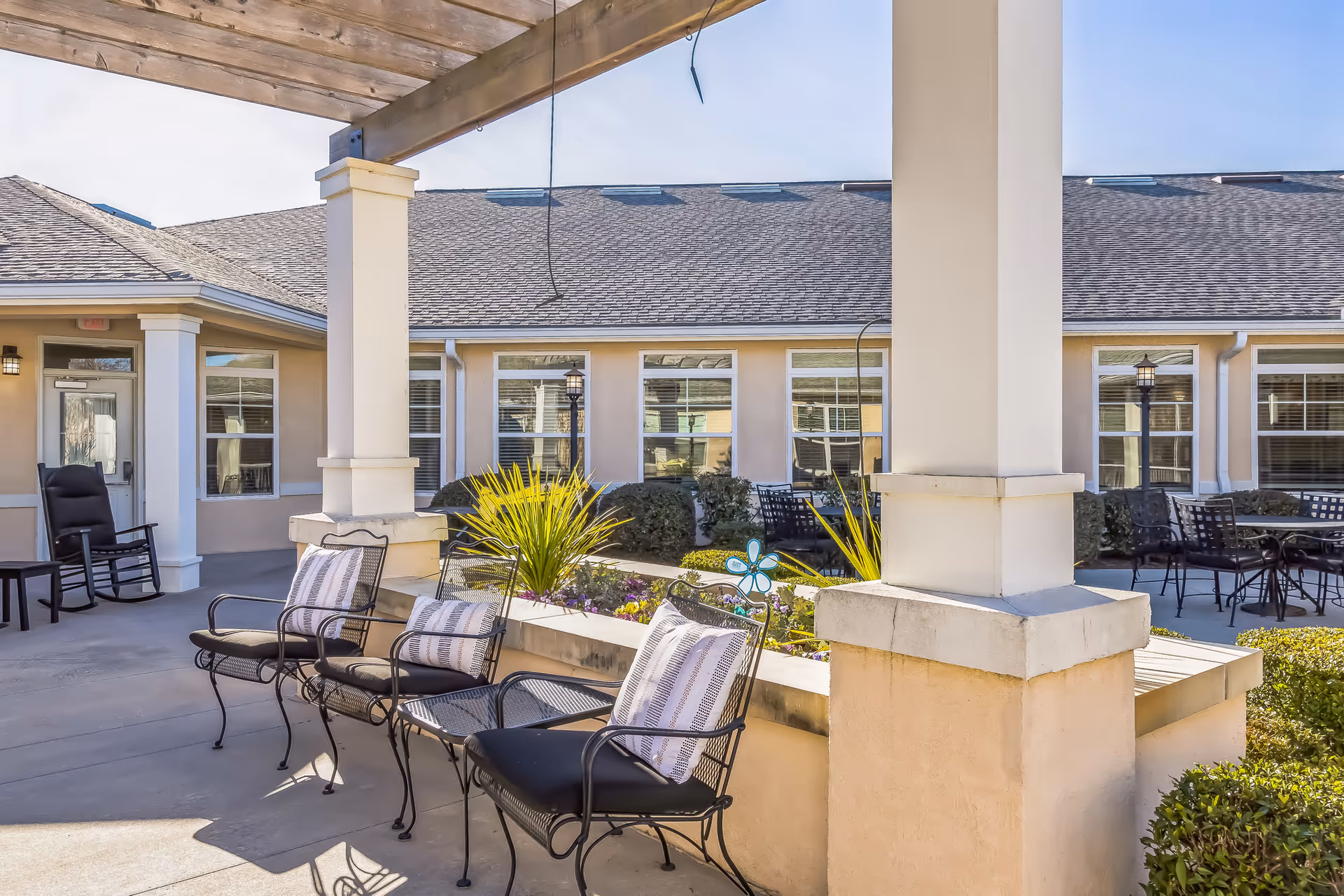Outdoor patio area at Barclay House of Augusta with metal chairs and cushions arranged around a planter with greenery and flowers, under a covered roof with columns. The building with multiple windows is visible in the background under a clear blue sky.