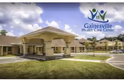 Exterior view of a single-story healthcare facility with a covered entrance and a driveway. The building is surrounded by a well-maintained lawn and trees under a partly cloudy sky. The logo and name 'Gainesville Health Care Center' are visible in the upper right corner.
