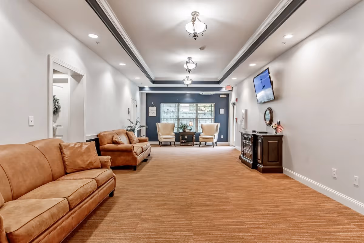 A spacious and well-lit living room area with two brown leather sofas on the left side, two beige armchairs near a window at the far end, and a dark wooden cabinet with a mounted flat-screen TV on the right wall. The room has beige carpet flooring, white walls, and ceiling lights with decorative fixtures.