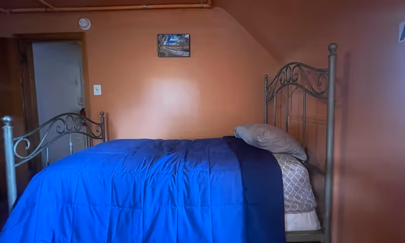 A bedroom with orange walls featuring a metal bed frame with a blue comforter and a pillow. There is a small framed picture hanging on the wall above the bed and a doorway leading to another room.