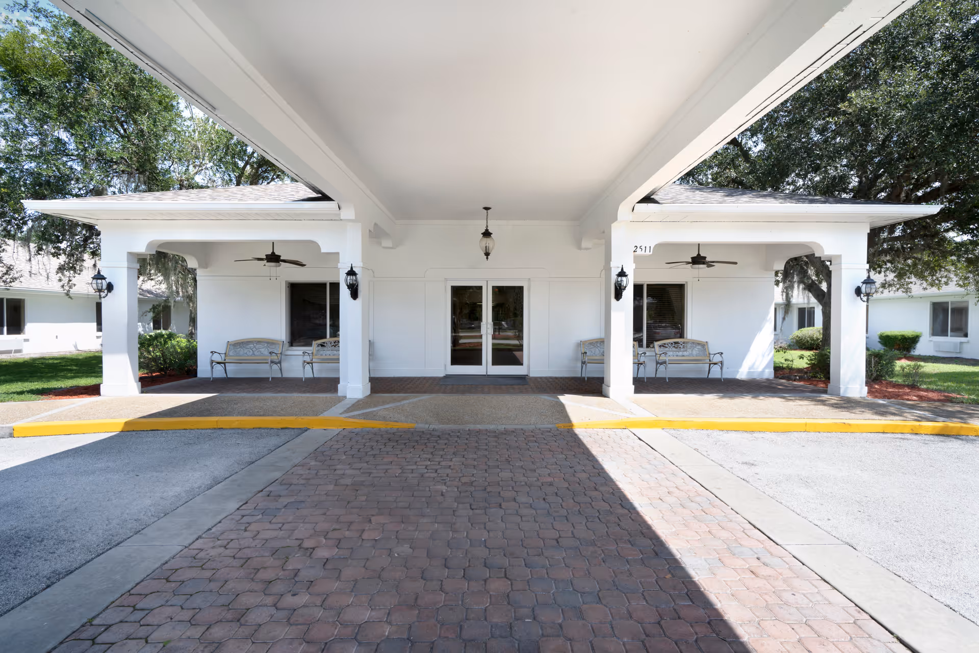 Entrance area of Harborview Health Center Kissimmee showing a covered driveway with two white pillars supporting the roof. There are benches on either side under the covered area, ceiling fans, and outdoor lantern-style lights mounted on the pillars. The building is white with windows and greenery around it.