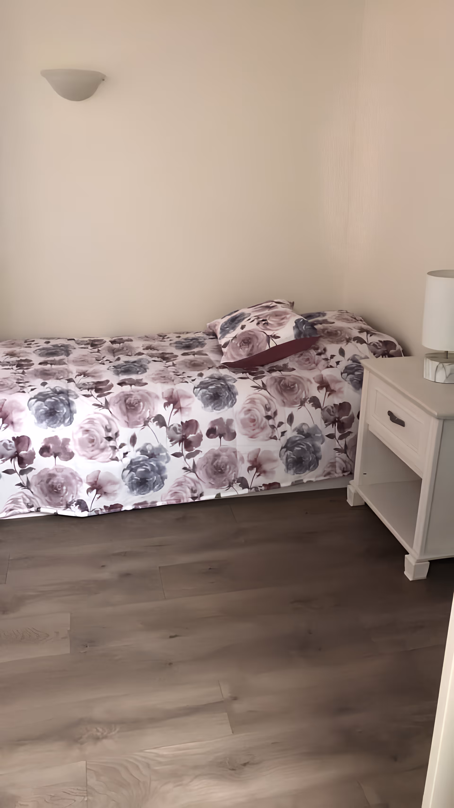 A simple bedroom with a single bed covered in a floral-patterned bedspread in shades of purple and gray. There is a matching pillow on the bed. Next to the bed is a white nightstand with a drawer and an open shelf, topped with a white lamp. The walls are plain and light-colored, and the floor is wooden.