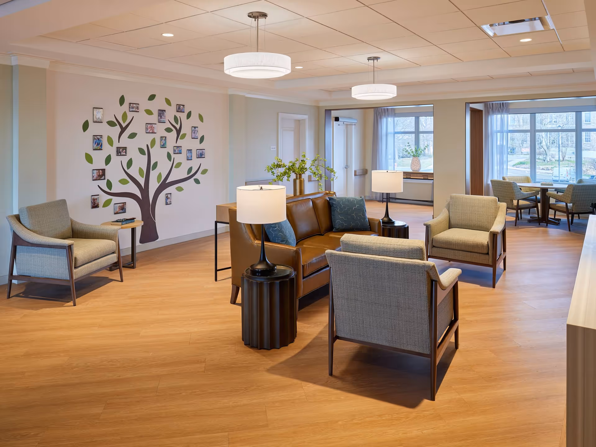 A bright and spacious living room area in The Wartburg Home featuring a brown leather sofa with blue pillows, two beige armchairs, and a small round black side table with a lamp. The room has light wood flooring, a wall decorated with a tree mural and framed photos, and large windows letting in natural light. In the background, there is a dining area with tables and chairs near the windows.