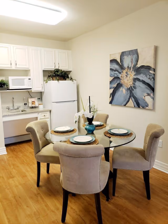 Small dining area with a round glass table set for four and upholstered chairs, with a kitchenette, white cabinets, and refrigerator in the background.