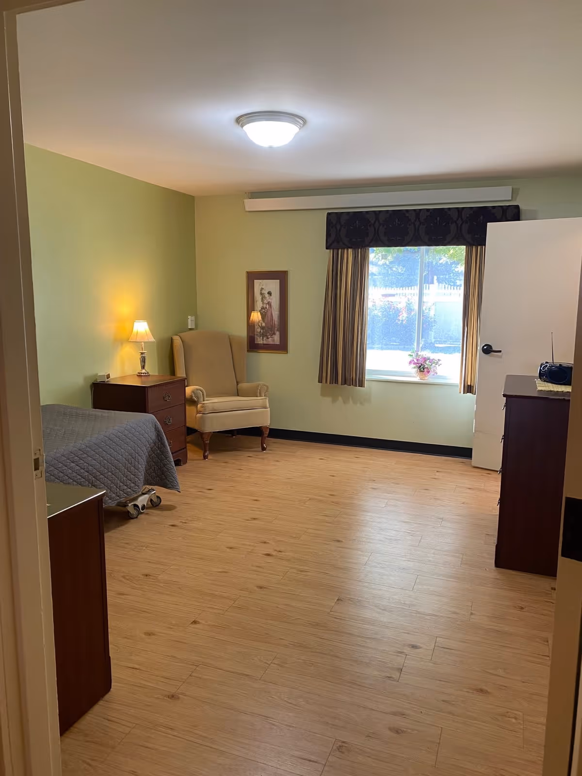 A simple, clean bedroom with light green walls and wood flooring. The room contains a single bed with a gray quilt, a wooden nightstand with a lamp, a beige upholstered armchair, a framed picture on the wall, a window with striped curtains and a valance, and a wooden dresser with a radio on top. The door to the room is open, letting in natural light.