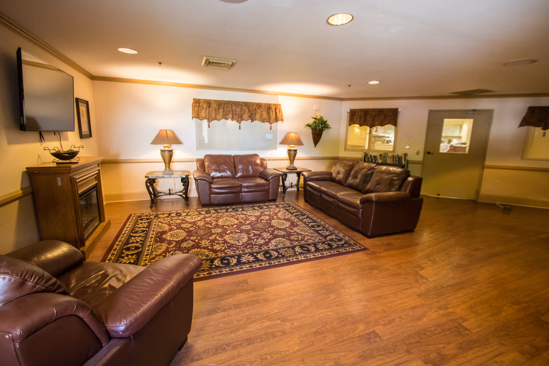 A cozy living room area in a nursing and rehabilitation center featuring three brown leather sofas arranged around a patterned area rug. There are two side tables with lamps, a wall-mounted TV above a wooden cabinet, and a window with a valance. The room has wooden flooring and soft lighting.
