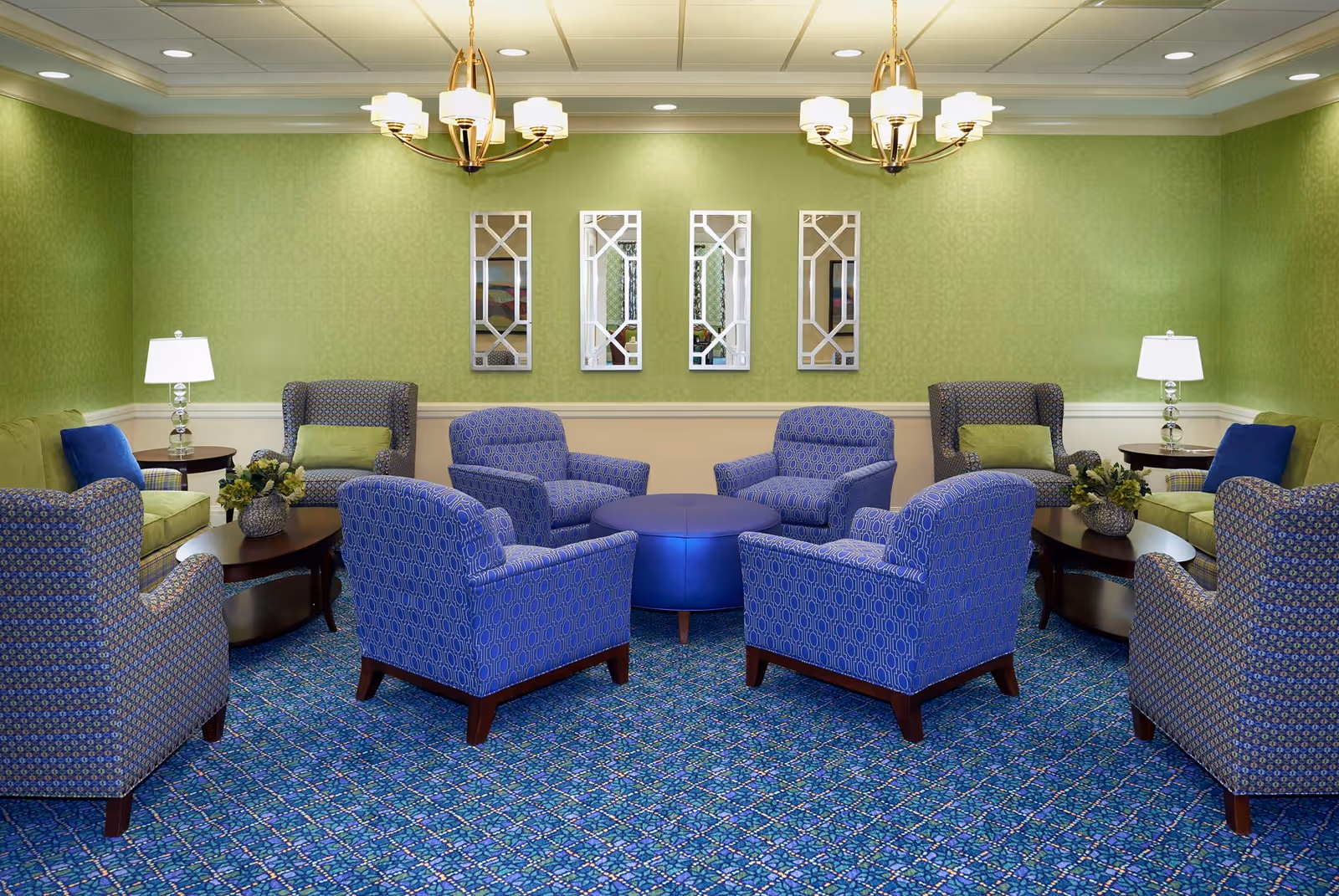 A cozy sitting area with a circular arrangement of eight blue patterned armchairs around a round blue ottoman. The room has green walls with four decorative mirrors hanging on one wall, two green sofas with blue pillows, two side tables with lamps, and two chandeliers hanging from the ceiling. The floor is covered with a blue patterned carpet.