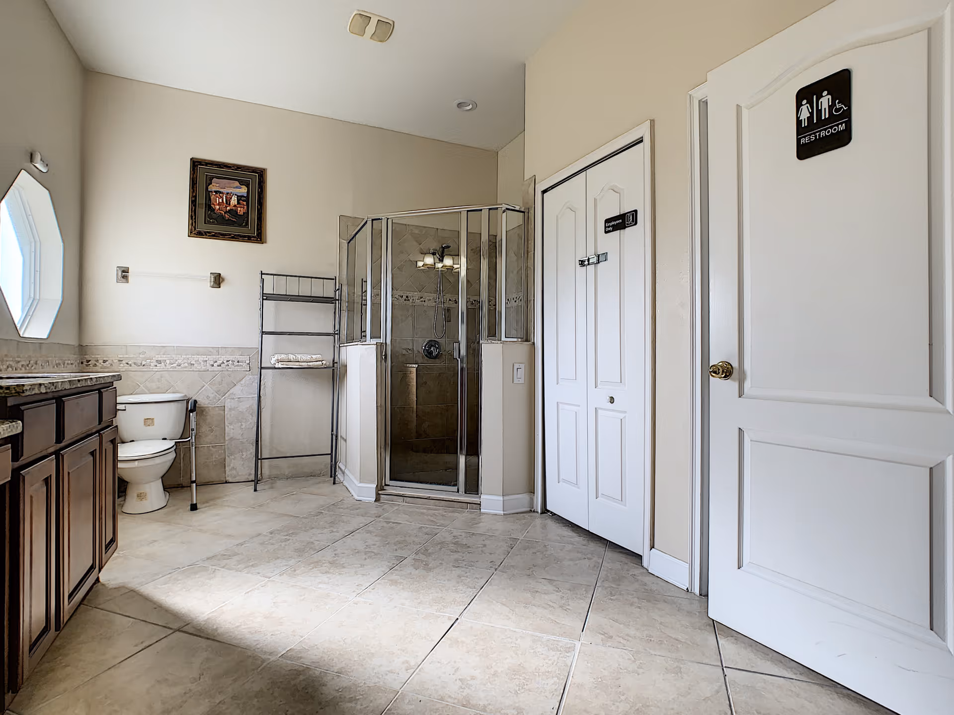 Bright tiled bathroom featuring a glass-enclosed shower, toilet, wood vanity, shelving unit, and a labeled restroom door.