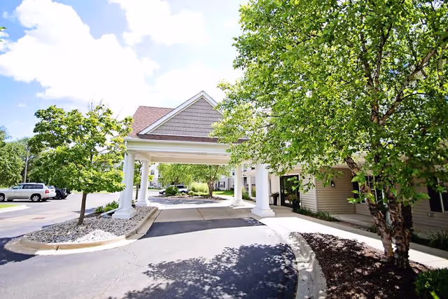 Entrance area of a senior living facility with a covered drop-off zone supported by white columns, surrounded by green trees and a parking lot with several cars under a partly cloudy sky.