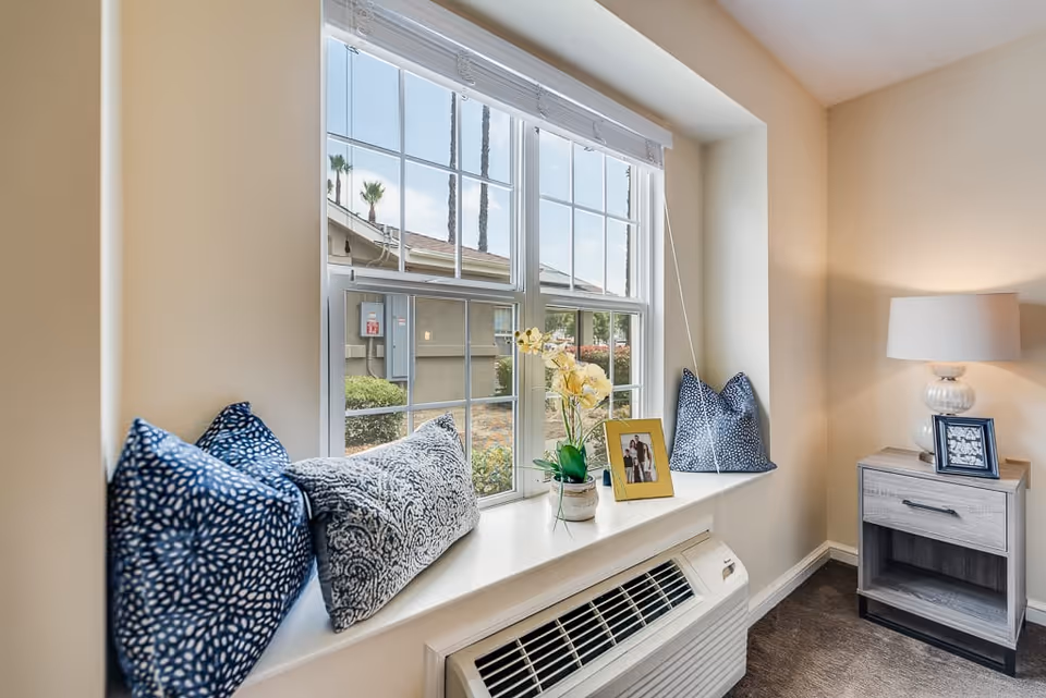 A cozy window nook with three decorative pillows and a potted orchid on the windowsill. Next to the window is a small wooden nightstand with a lamp and two framed photographs. Outside the window, some palm trees and a neighboring building are visible.
