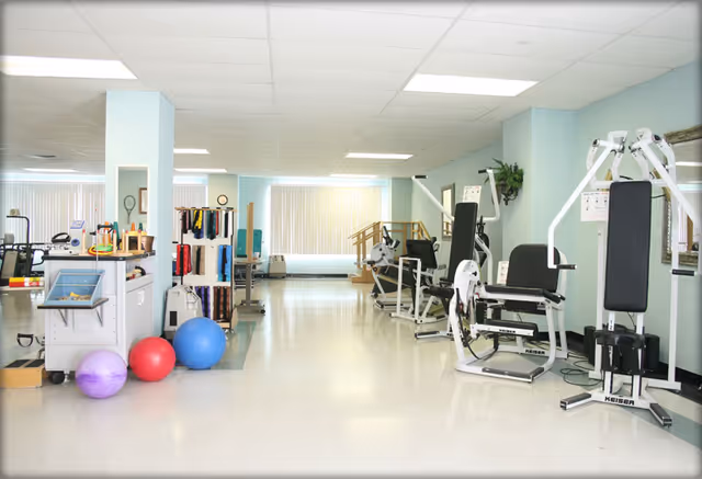 A bright and spacious physical therapy or rehabilitation room with exercise equipment including weight machines, therapy balls, and resistance bands. The room has light blue walls, a white ceiling with fluorescent lights, and large windows with vertical blinds letting in natural light.
