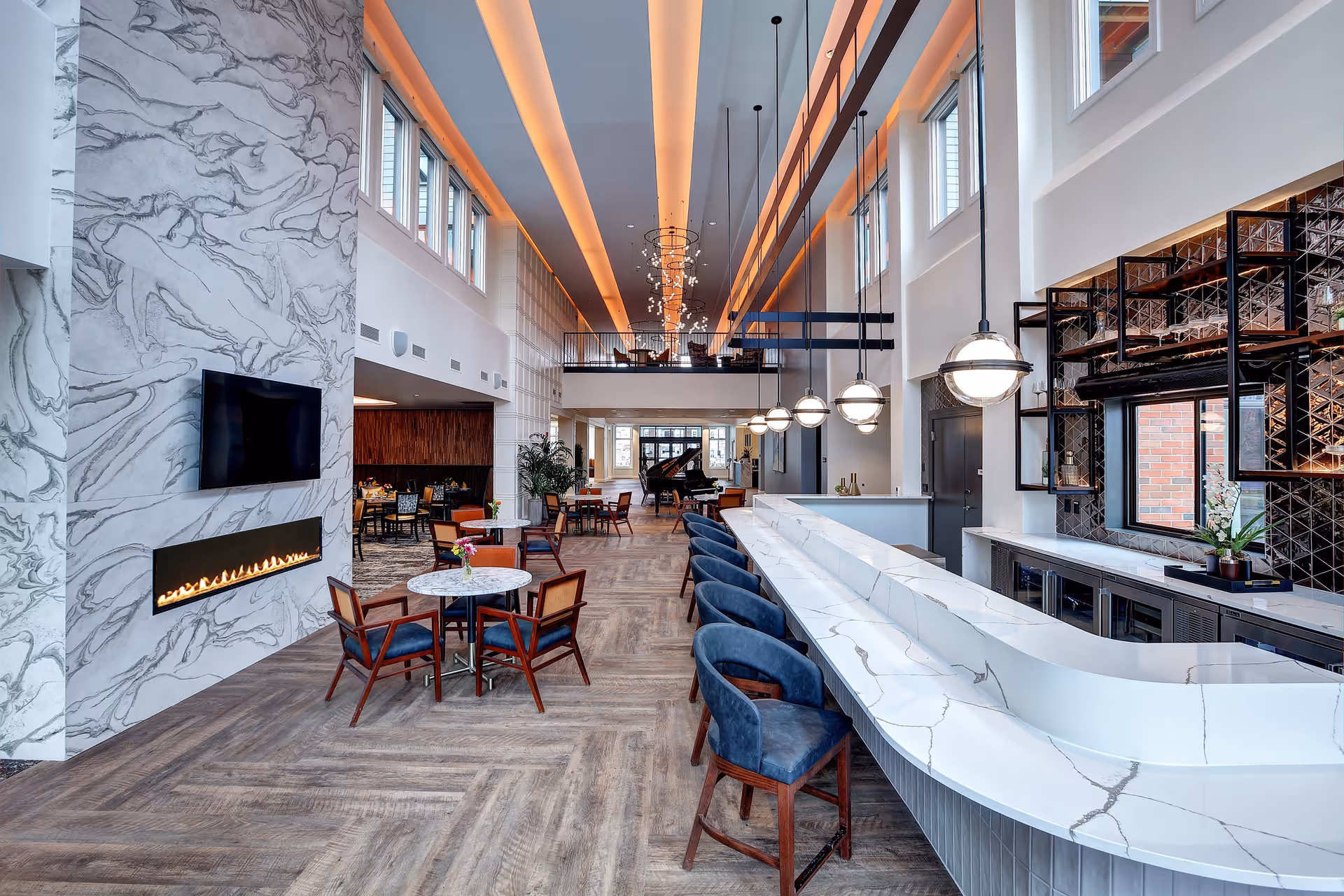 A spacious, modern common area in The Delaney at the Vale featuring a long white marble bar with blue cushioned bar stools, round tables with chairs, a wall-mounted TV above a linear fireplace with a marble surround, large windows, and a grand piano in the background under high ceilings with ambient lighting.
