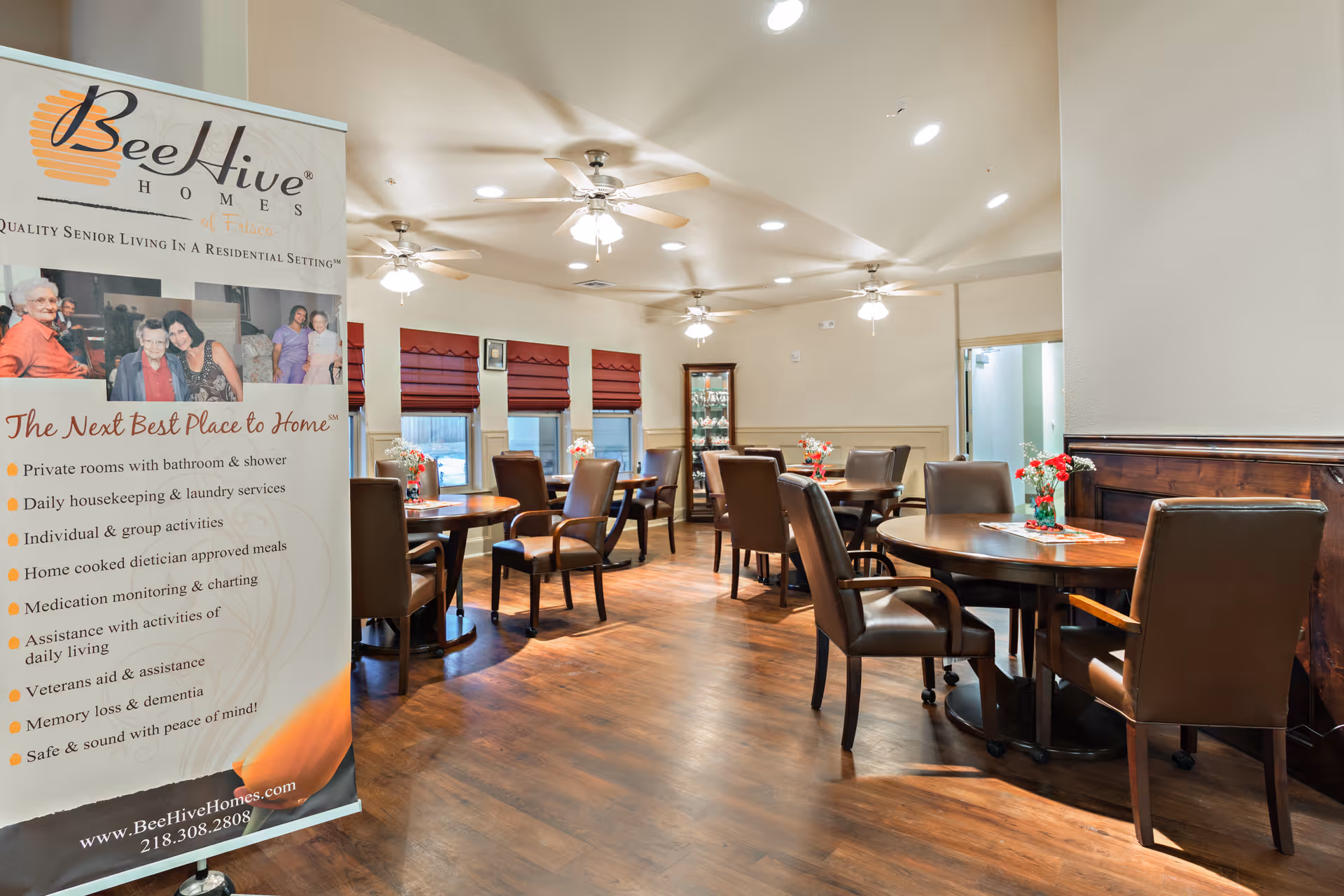 Bright communal dining room with round tables, leather chairs, ceiling fans, and a BeeHive Homes promotional banner on the left.