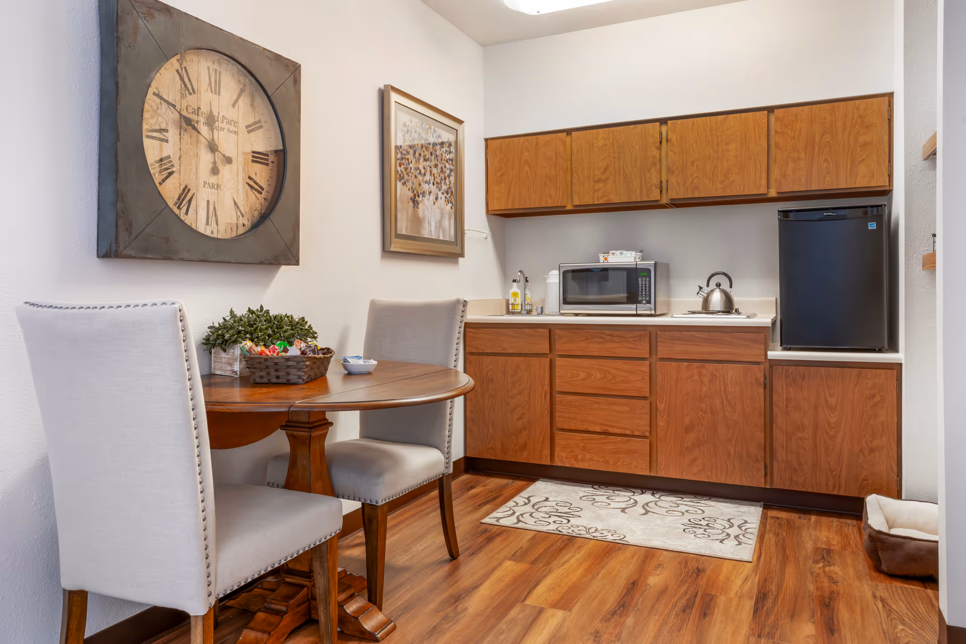 Small kitchenette and dining nook with a round wooden table, two upholstered chairs, a large wall clock, microwave, mini fridge, and wooden cabinets.