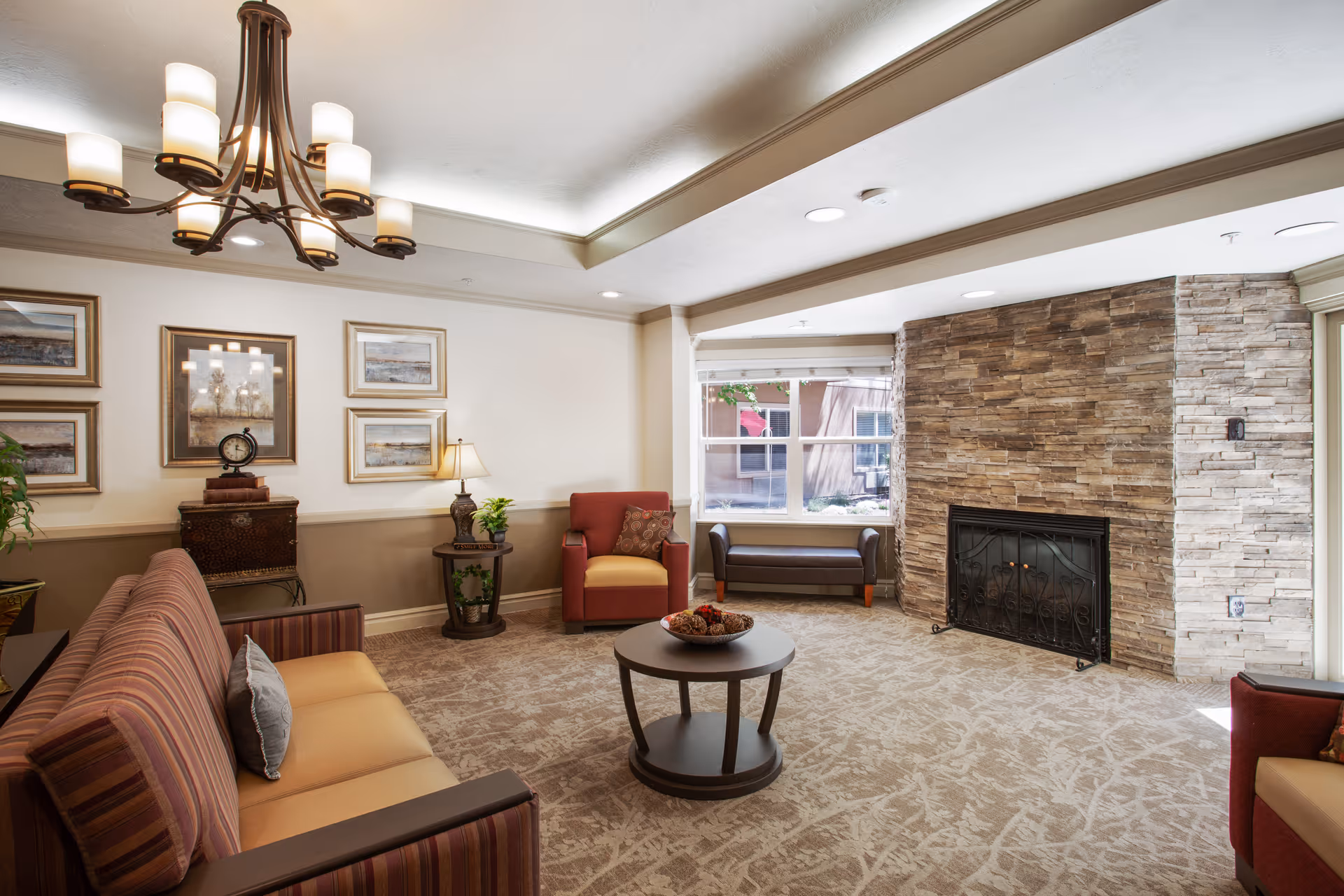 A cozy living room area with a striped sofa, a red armchair, a round coffee table with decorative items, a stone fireplace, a window with a bench underneath, framed artwork on the wall, and a chandelier hanging from the ceiling.