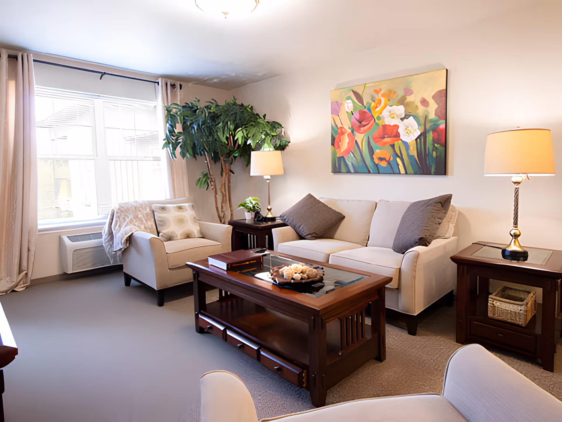 A cozy living room with a beige sofa and armchair, a wooden coffee table with decorative items, two side tables with lamps, a large leafy plant near the window, and a colorful floral painting on the wall.