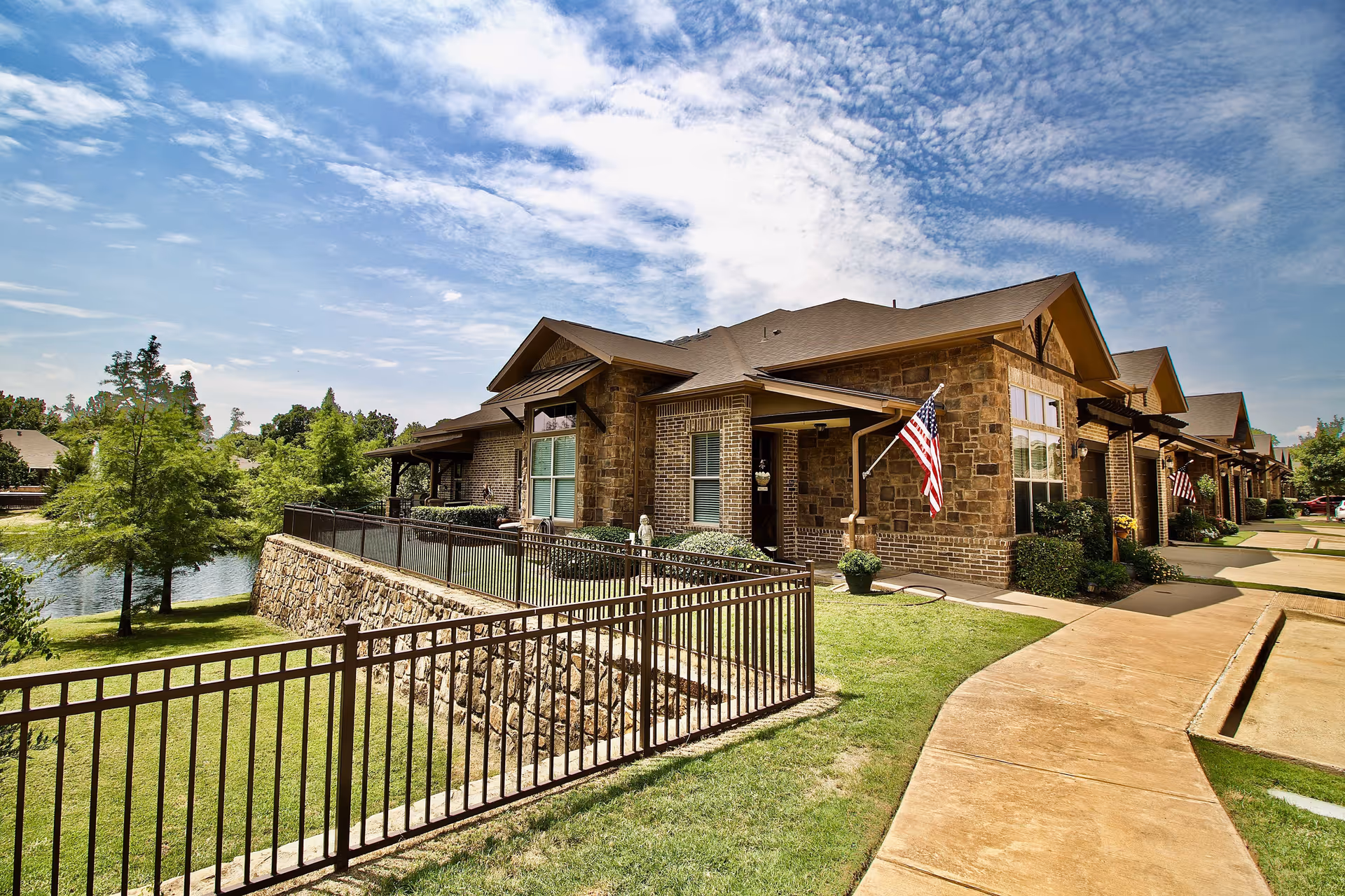 Exterior view of a senior living facility with stone and brick buildings, a paved walkway, green lawns, and an American flag displayed near the entrance. Trees and a small body of water are visible in the background under a partly cloudy sky.