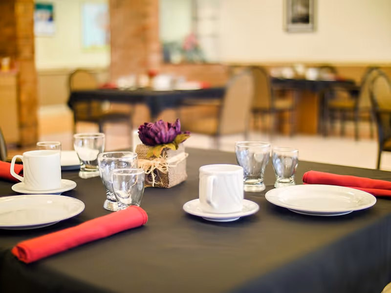 A dining table set with white plates, cups, glasses, and red cloth napkins on a black tablecloth. A small decorative purple flower centerpiece is placed in the middle of the table. In the background, there are more tables and chairs in a softly lit dining area.