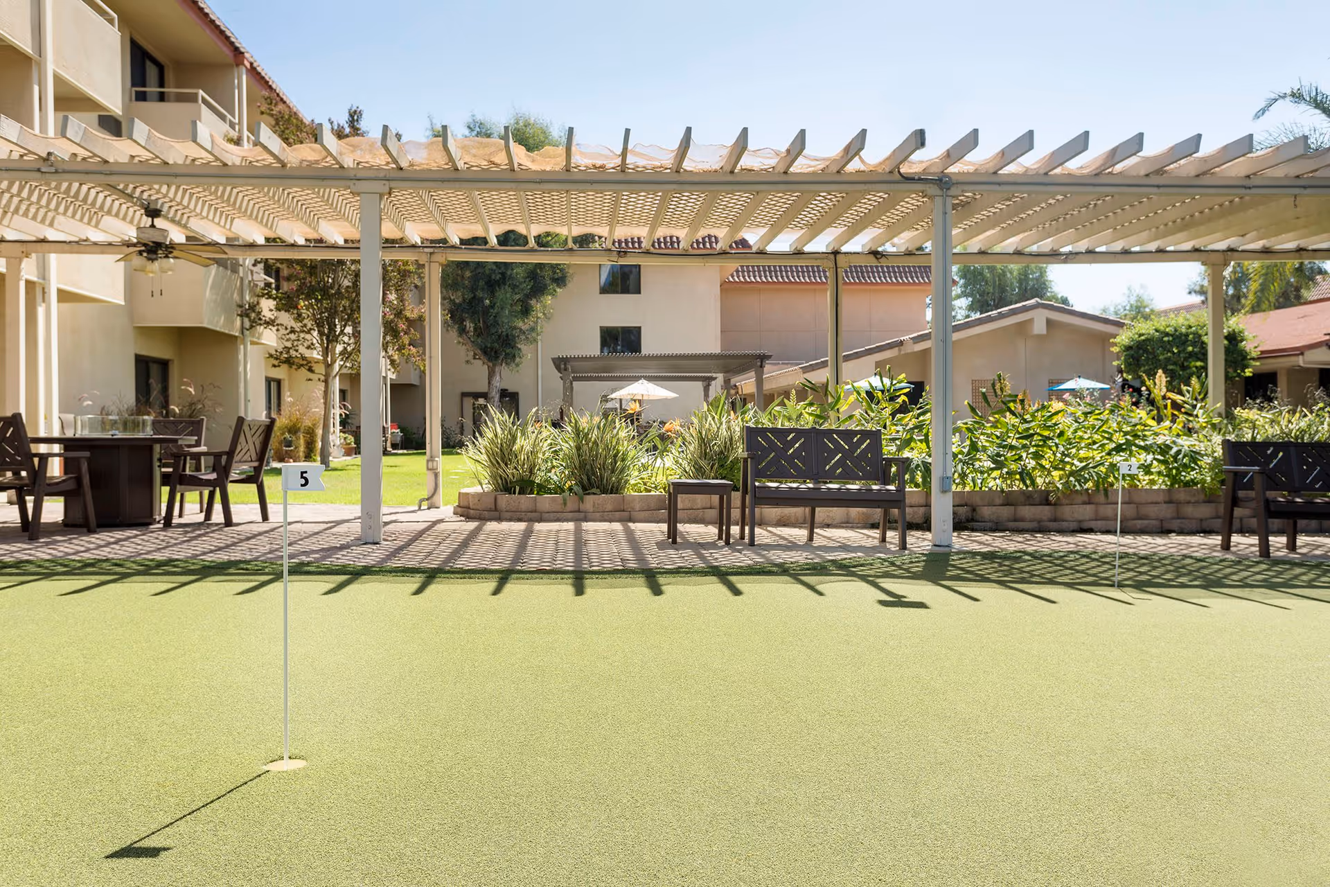 Outdoor putting green area with two small flags numbered 5 and 2, shaded seating with benches and chairs under a pergola, surrounded by greenery and buildings in the background.