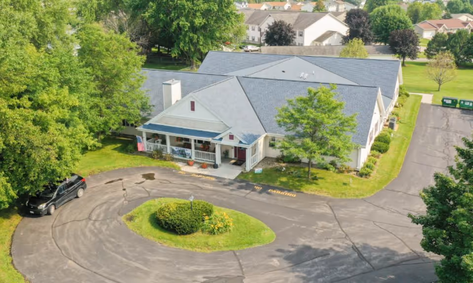 Aerial view of the front of a single-story assisted living building with a circular driveway, lawn and surrounding trees.