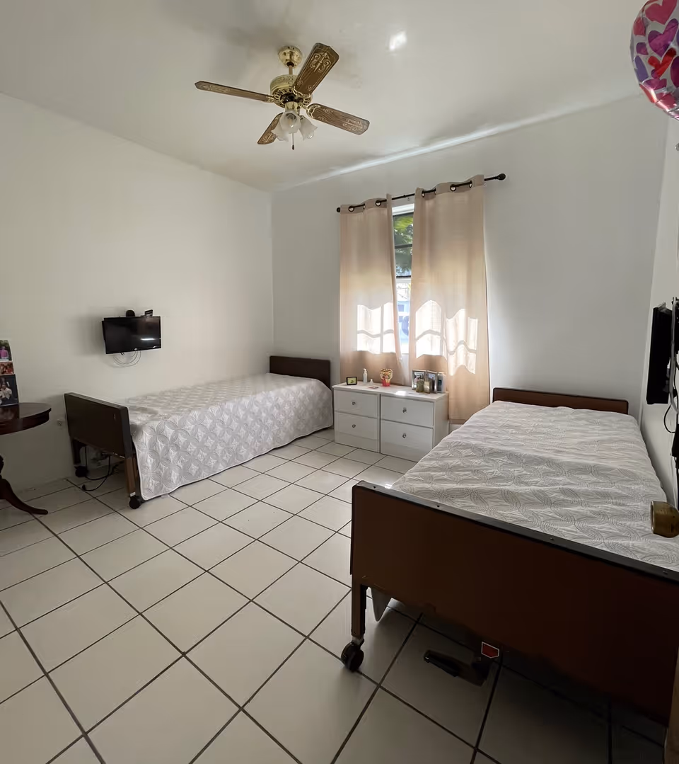 A simple bedroom with two single beds covered with white patterned bedspreads. Between the beds is a white dresser with personal items and framed photos on top. A window with beige curtains lets in natural light. A small wall-mounted TV is on the left wall, and a ceiling fan with wooden blades is overhead. The floor is tiled with large white tiles.