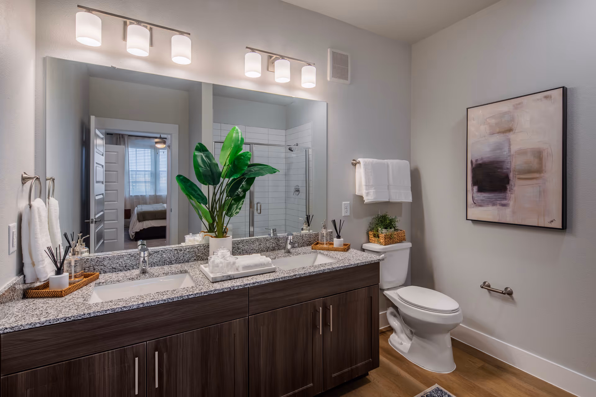 Modern bathroom with a double sink vanity featuring granite countertops and dark wood cabinets. Above the sinks is a large mirror with six light fixtures. A green potted plant and neatly arranged toiletries are on the counter. A white toilet is next to the vanity with a towel rack above it holding two white towels. A framed abstract painting hangs on the wall. The bathroom has light-colored walls and wood flooring. A doorway leads to a bedroom with a bed and window visible.