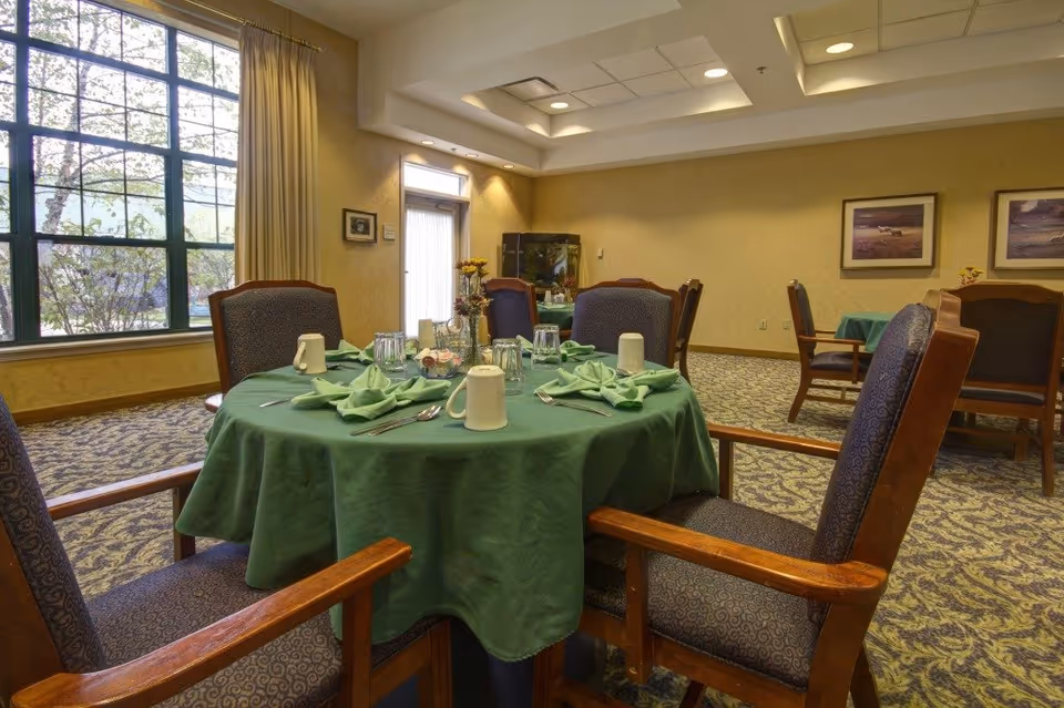A dining room in an assisted living facility with round tables covered in green tablecloths, set with folded green napkins, cups, glasses, and silverware. The room has patterned carpet, large windows with beige curtains, framed pictures on the walls, and wooden chairs with cushioned seats and backs.