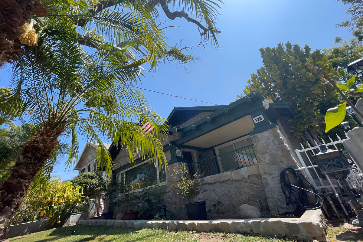 Front exterior of a stone-faced bungalow with a covered porch, palm trees in the yard, and an American flag.