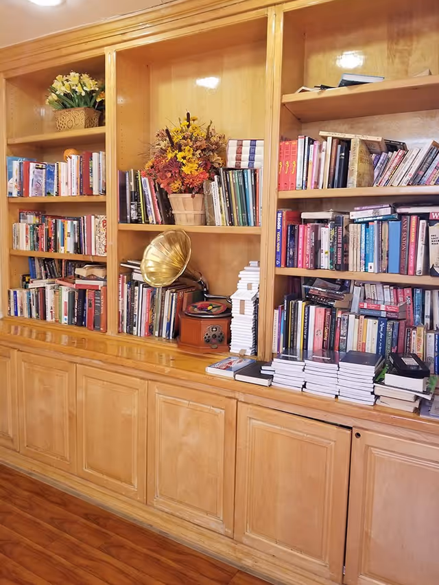 Wooden bookshelf filled with numerous books, a vintage gramophone, and decorative flower arrangements in a cozy indoor setting with wooden flooring.