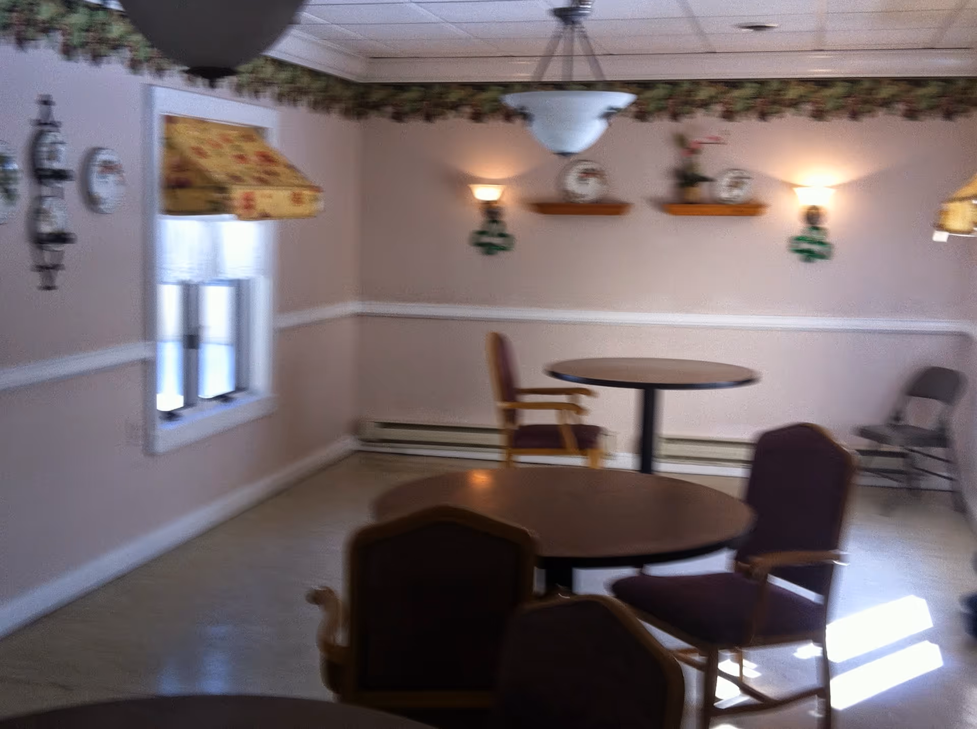 A small dining area with round tables and chairs, floral valances on the windows, wall-mounted shelves with decorative plates and a small plant, and soft wall lighting fixtures.