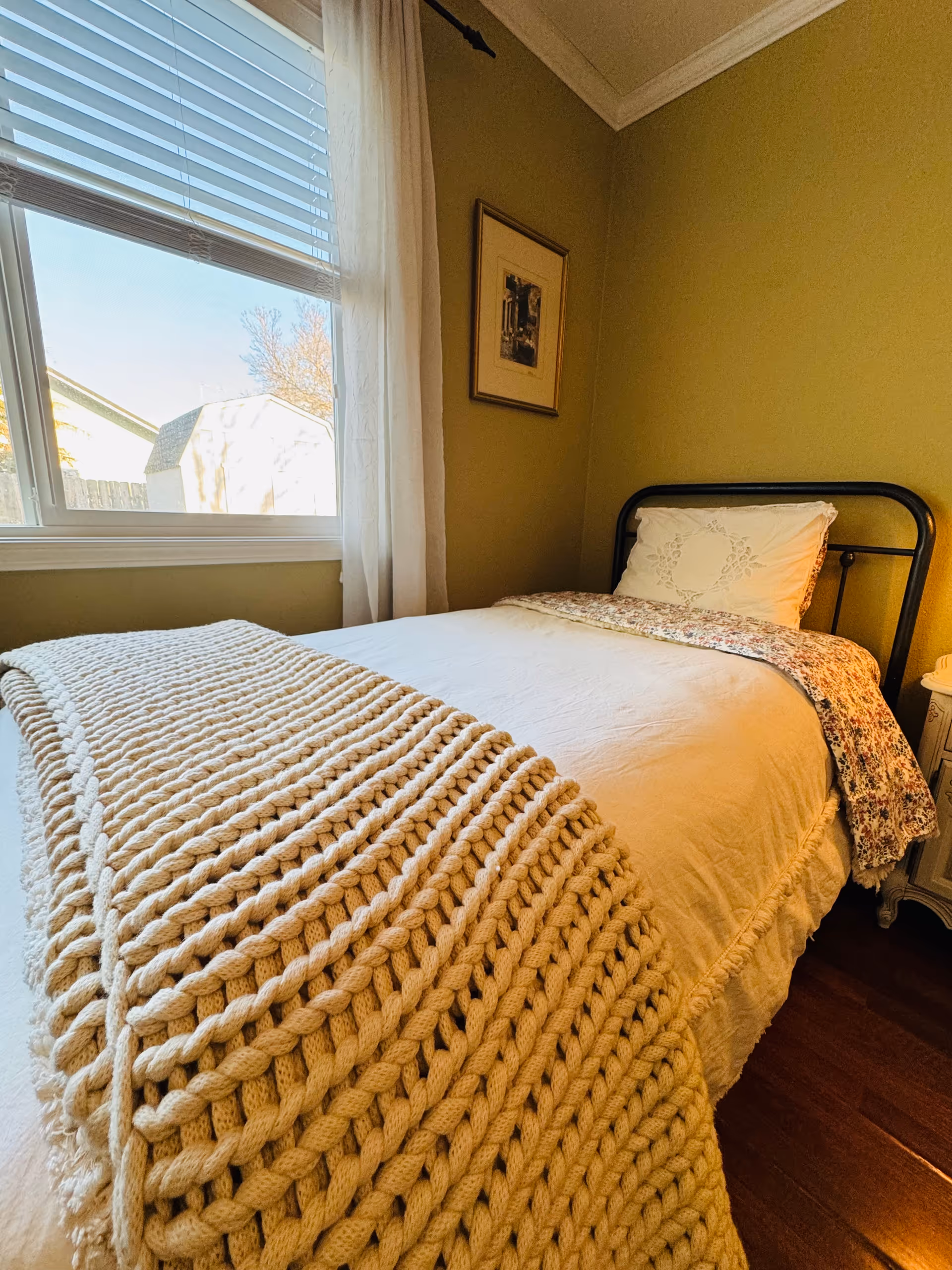 A cozy bedroom with a single bed featuring a black metal frame, white bedding, a floral blanket, and a chunky knit beige throw. There is a window with white blinds and sheer curtains letting in natural light, a framed picture on the olive-green wall, and a small white nightstand beside the bed.