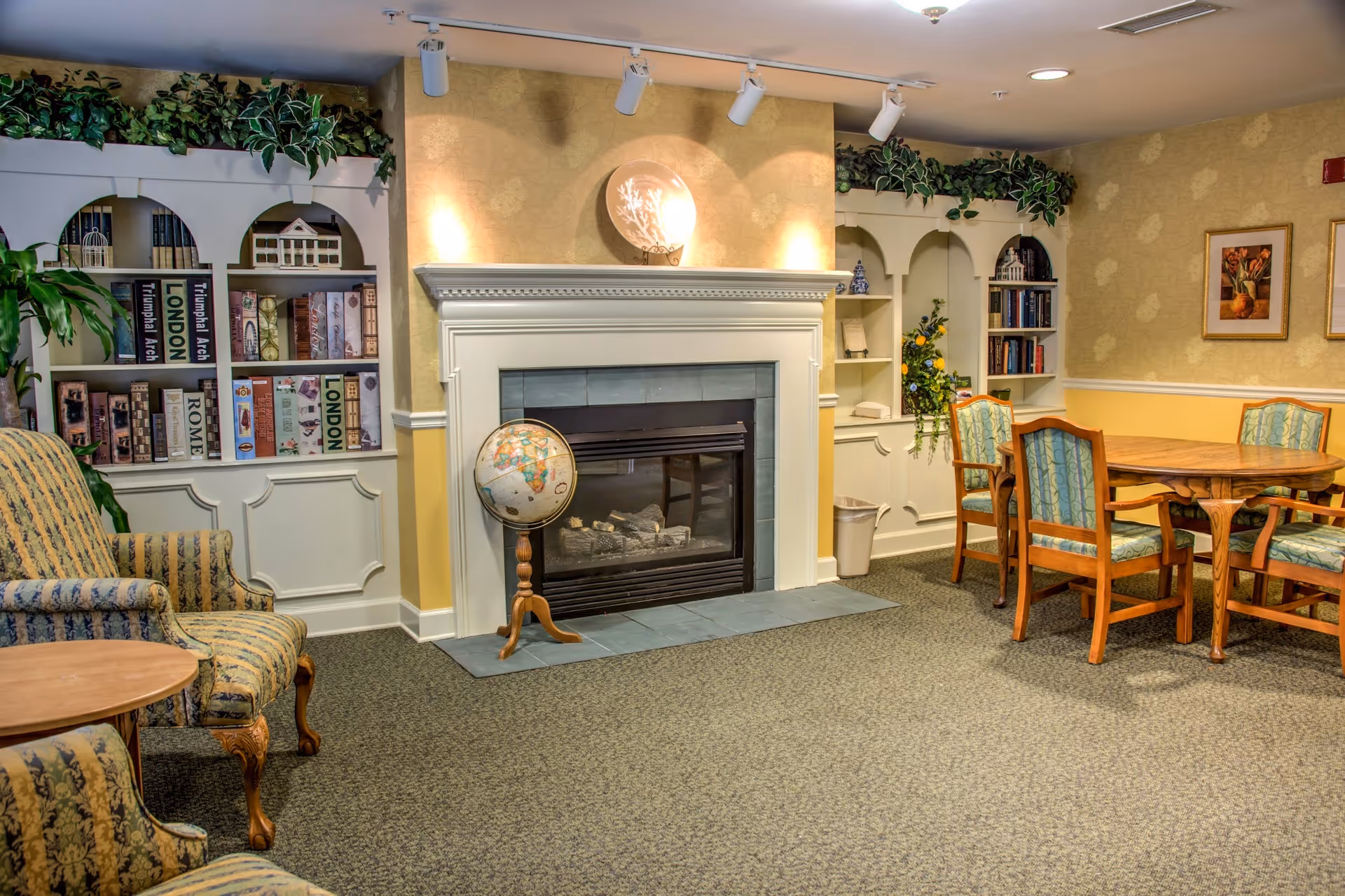 A cozy living room area with a fireplace in the center, flanked by built-in white bookshelves filled with books and decorative items. There is a globe on a wooden stand in front of the fireplace. To the right, there is a wooden table with four upholstered chairs. On the left side, there are two patterned armchairs and a small round wooden table. The walls are decorated with framed artwork and greenery above the bookshelves.
