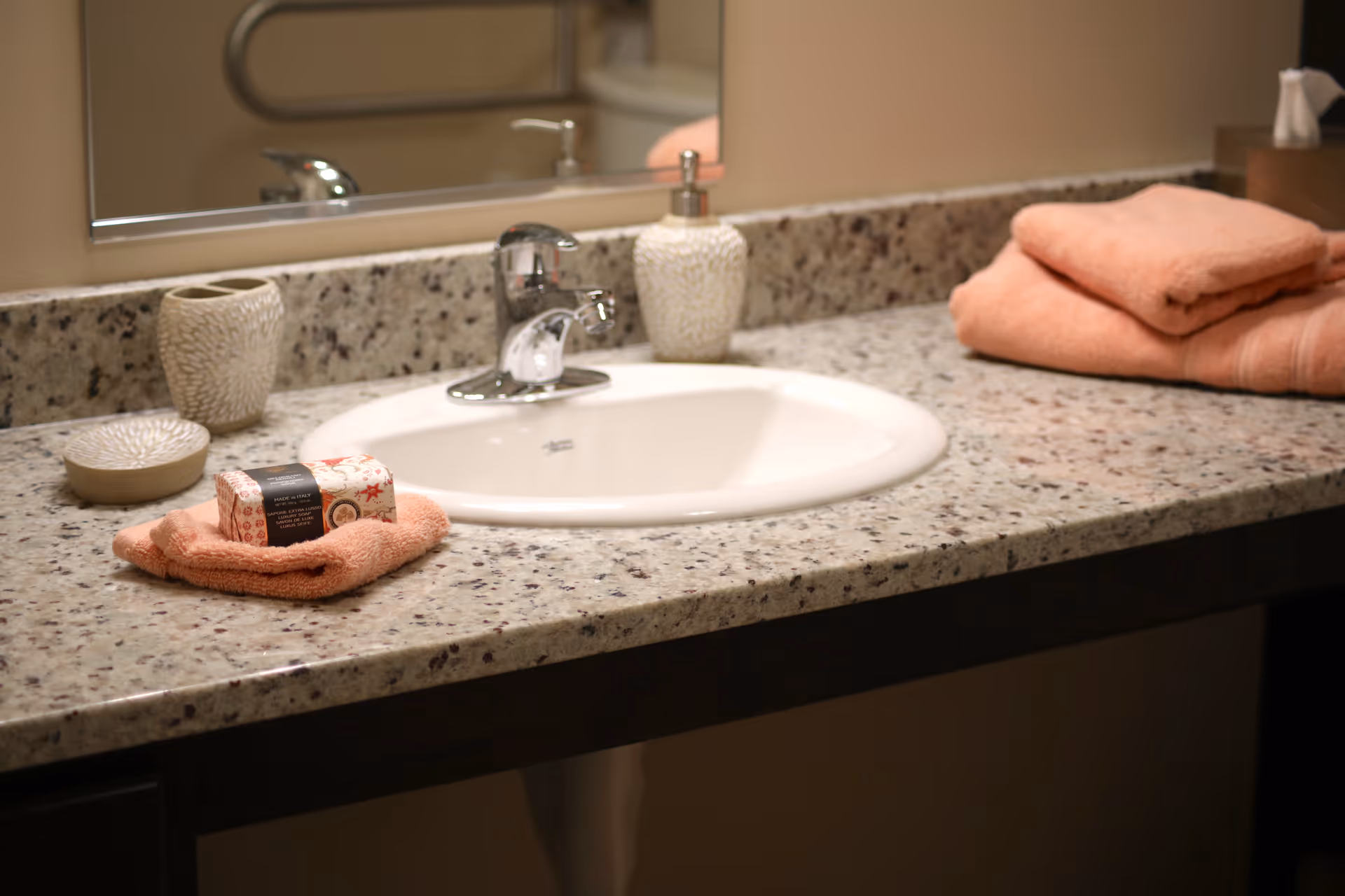 A bathroom sink with a granite countertop. On the countertop are a soap dispenser, a toothbrush holder, a small dish, a folded peach-colored washcloth with a bar of soap on top, and a stack of folded peach-colored towels. A mirror is visible behind the sink.