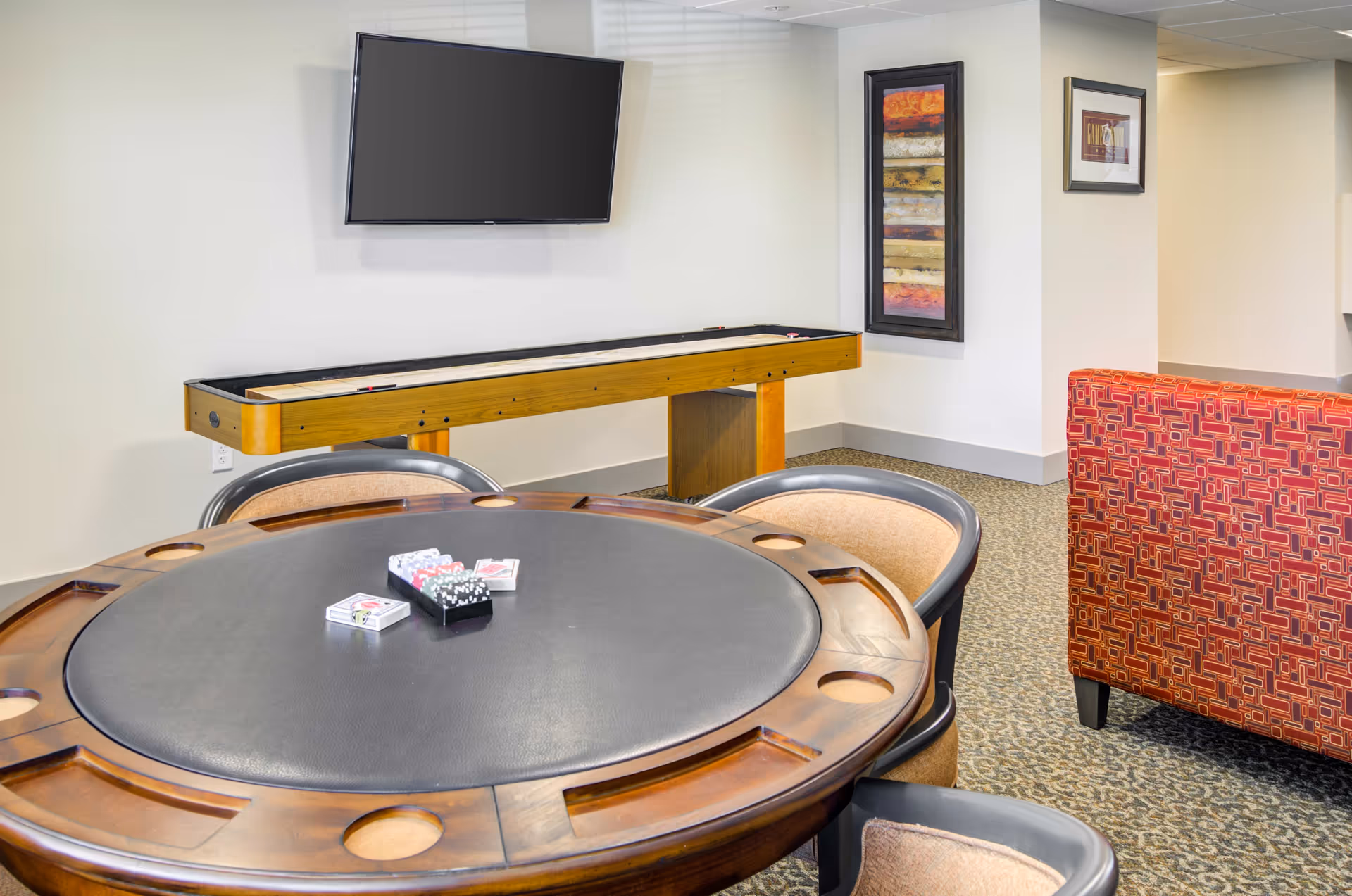 A game room with a round poker table featuring card decks and poker chips, surrounded by chairs. In the background, there is a shuffleboard table and a wall-mounted flat-screen TV. The room has carpeted flooring, light-colored walls, and framed artwork on the walls. A red patterned sofa is partially visible on the right side.