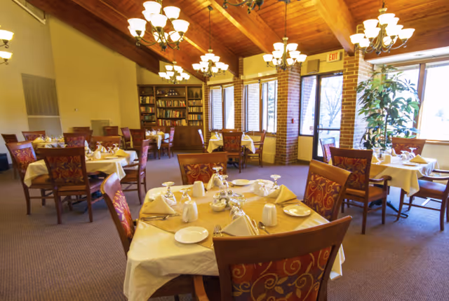 Dining room with tables set for meals, patterned chairs, chandeliers, and a vaulted wood-beamed ceiling.