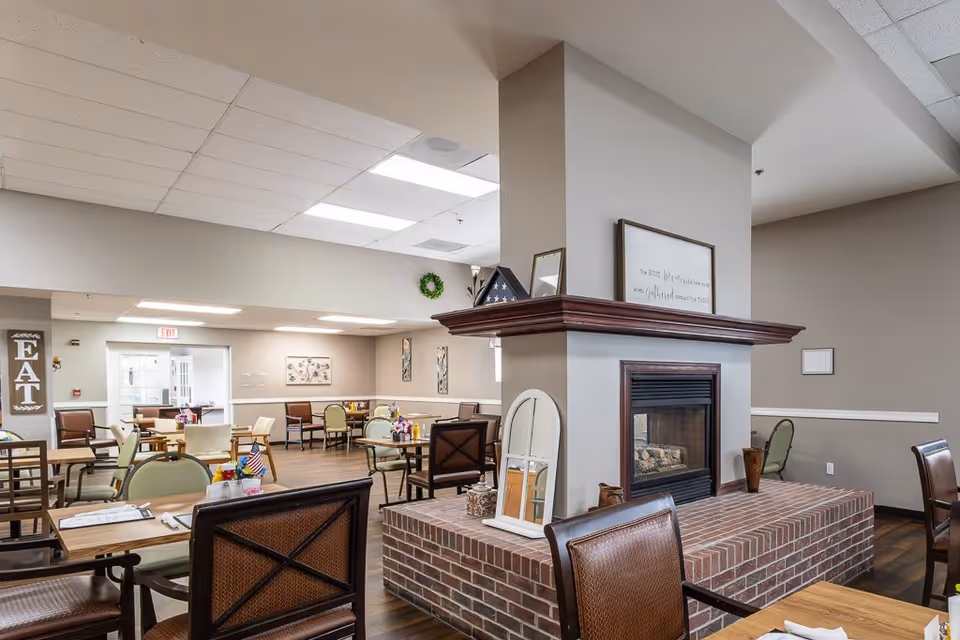 A spacious dining room in a senior living facility with multiple tables and chairs arranged for meals. The room features a central brick fireplace with a wooden mantle, decorated with framed pictures and a folded American flag. The walls are painted beige, and there are various wall decorations including a sign that says 'EAT'. The ceiling has fluorescent lighting, and the floor is wood.