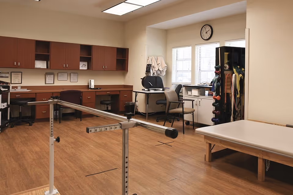 Therapy room with parallel bars, treatment table, cabinets, chairs and exercise equipment.