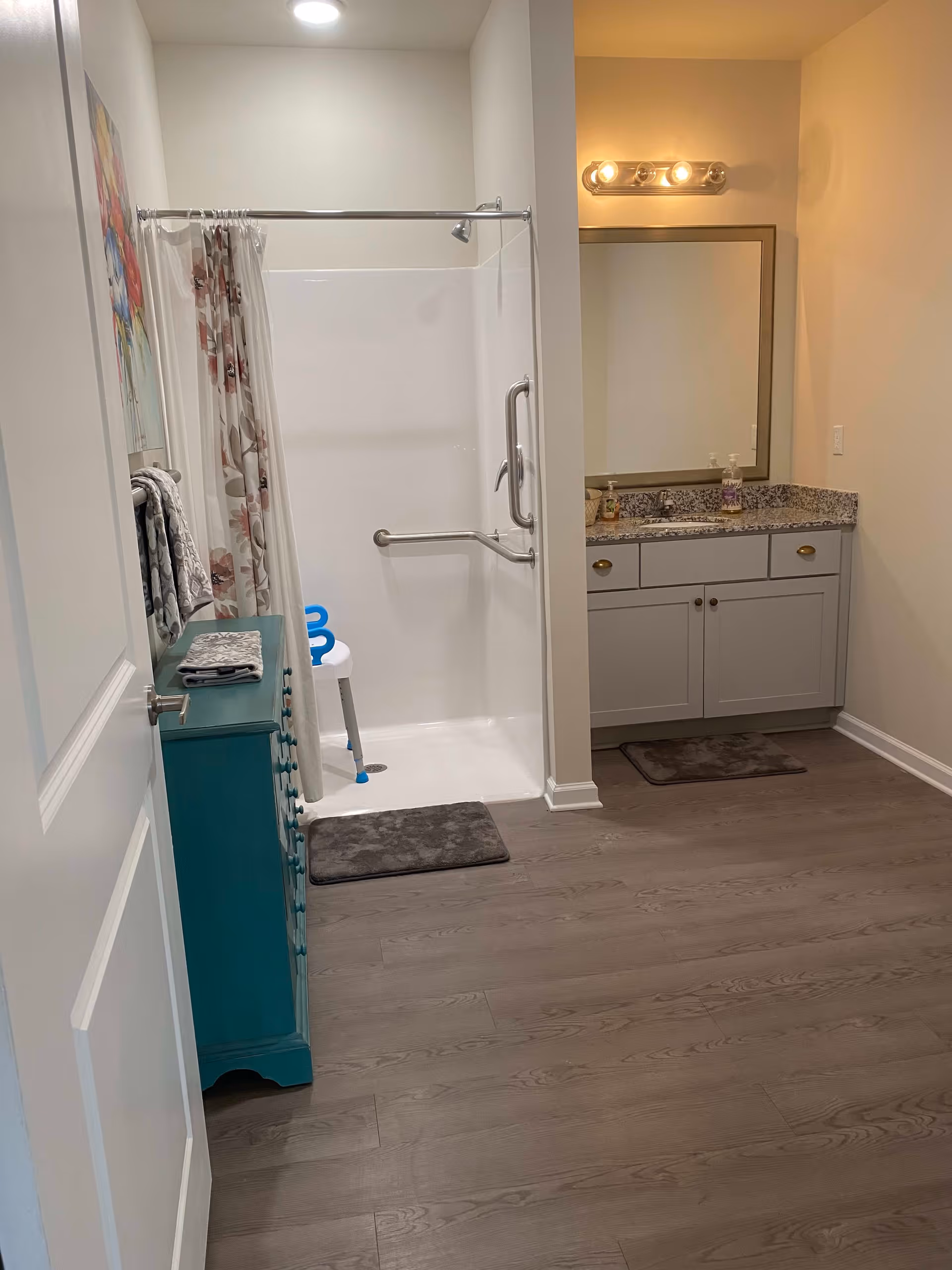A bathroom with a walk-in shower featuring a grab bar and a white shower chair with blue handles. There is a gray bath mat in front of the shower and another in front of a vanity with a granite countertop, sink, and large mirror. A teal cabinet with folded towels on top is positioned near the door. The floor is wood-style laminate, and the walls are painted light beige.