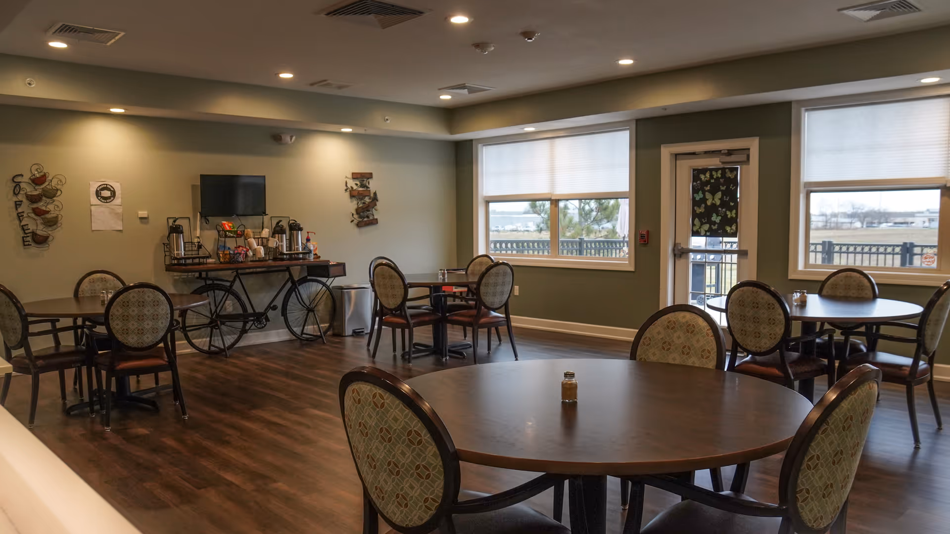 Spacious communal dining room with round tables and chairs, a coffee station against the wall, and windows with an exterior door.