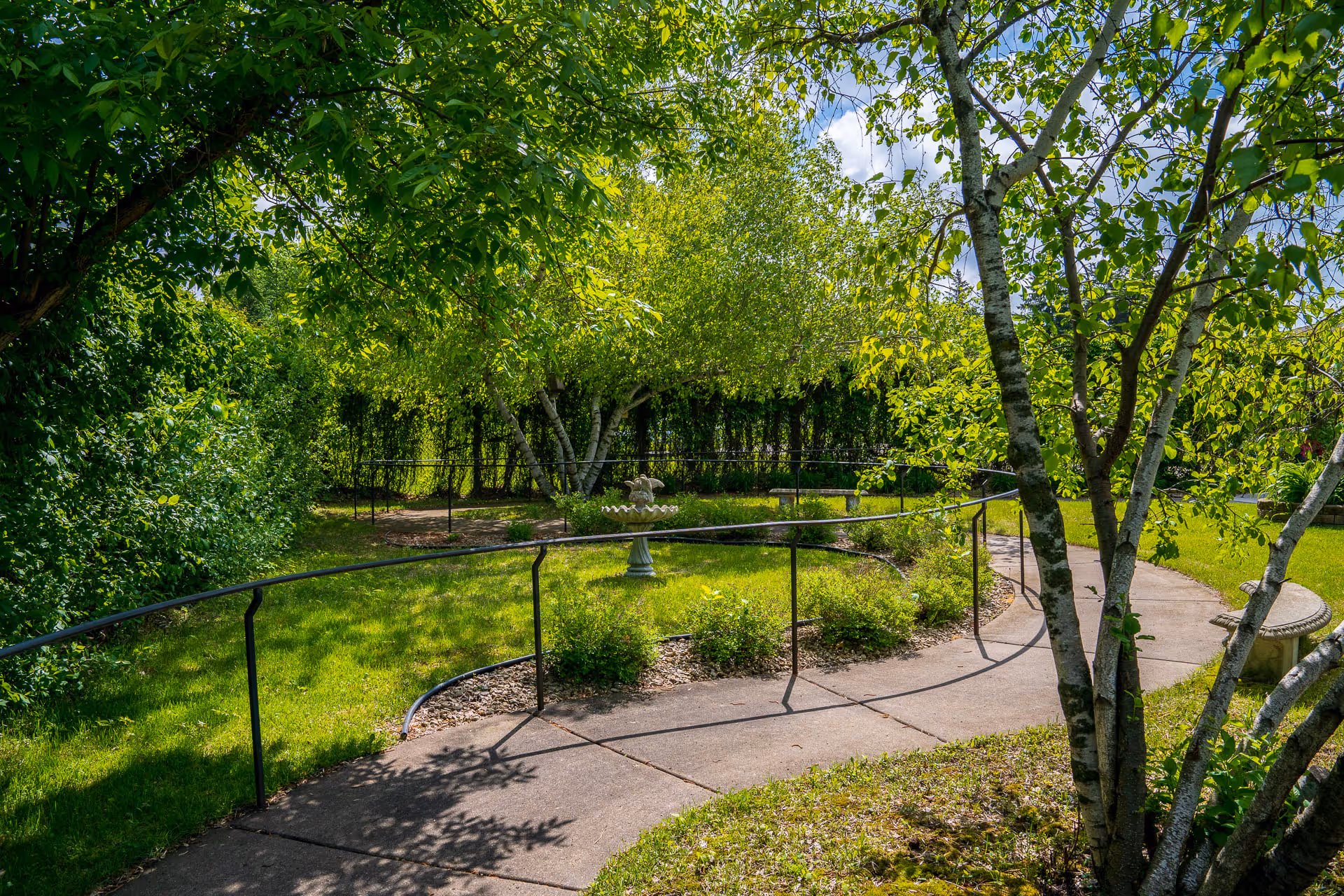 Curving paved garden path with a metal handrail surrounded by green trees, shrubs, a birdbath and benches.