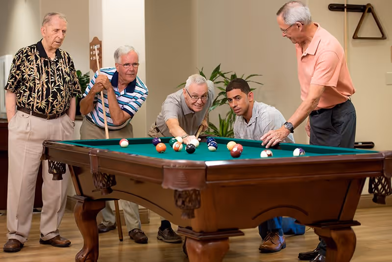 Five elderly men playing pool indoors, with one man aiming to strike the cue ball while the others watch and assist around the pool table.