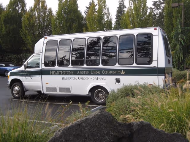 A white shuttle bus with green stripes parked in a lot surrounded by greenery and trees. The bus has the text 'Hearthstone Assisted Living Community, Beaverton, Oregon, 641-0911' written on its side.