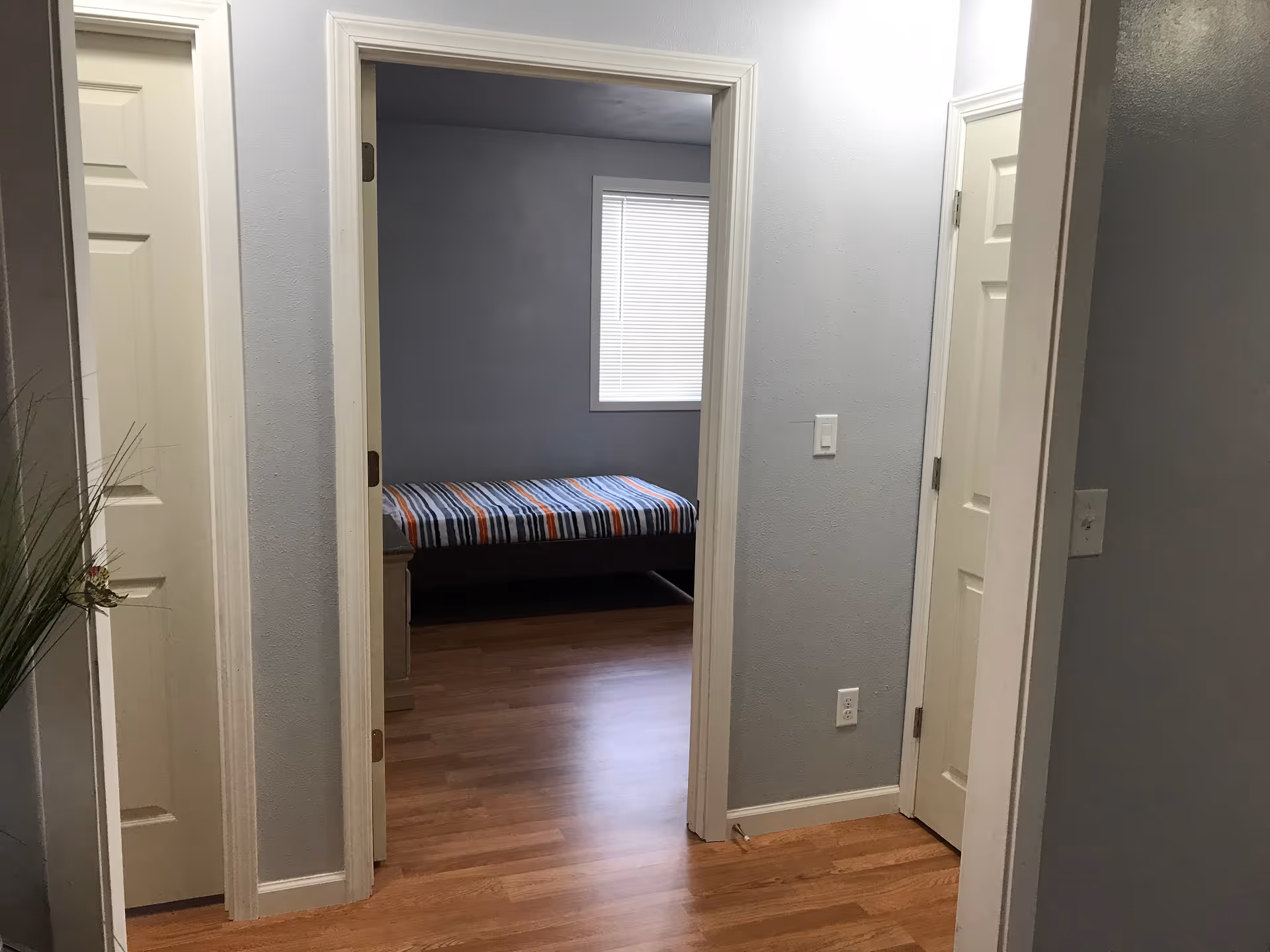 View of a hallway with light gray walls and wooden flooring leading to a bedroom with a bed covered in a striped blanket. There are two closed white doors on either side of the hallway and a window with blinds in the bedroom.