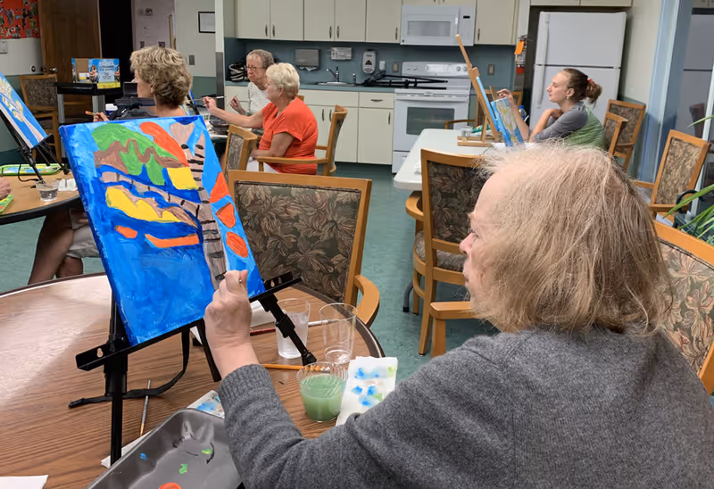 Several elderly individuals seated around tables in a room with kitchen cabinets and appliances, engaged in painting colorful pictures on canvases set on easels.