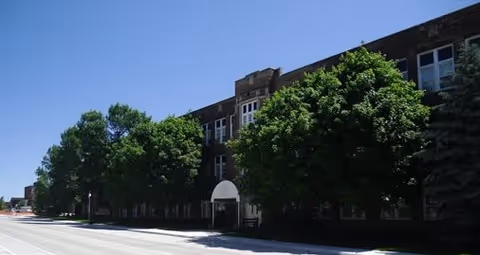 Exterior view of a multi-story brick building partially obscured by large green trees under a clear blue sky.