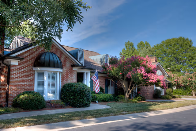 Exterior view of a brick building with white-trimmed windows, an American flag near the entrance, and landscaped greenery including bushes and a tree with pink flowers under a clear blue sky.