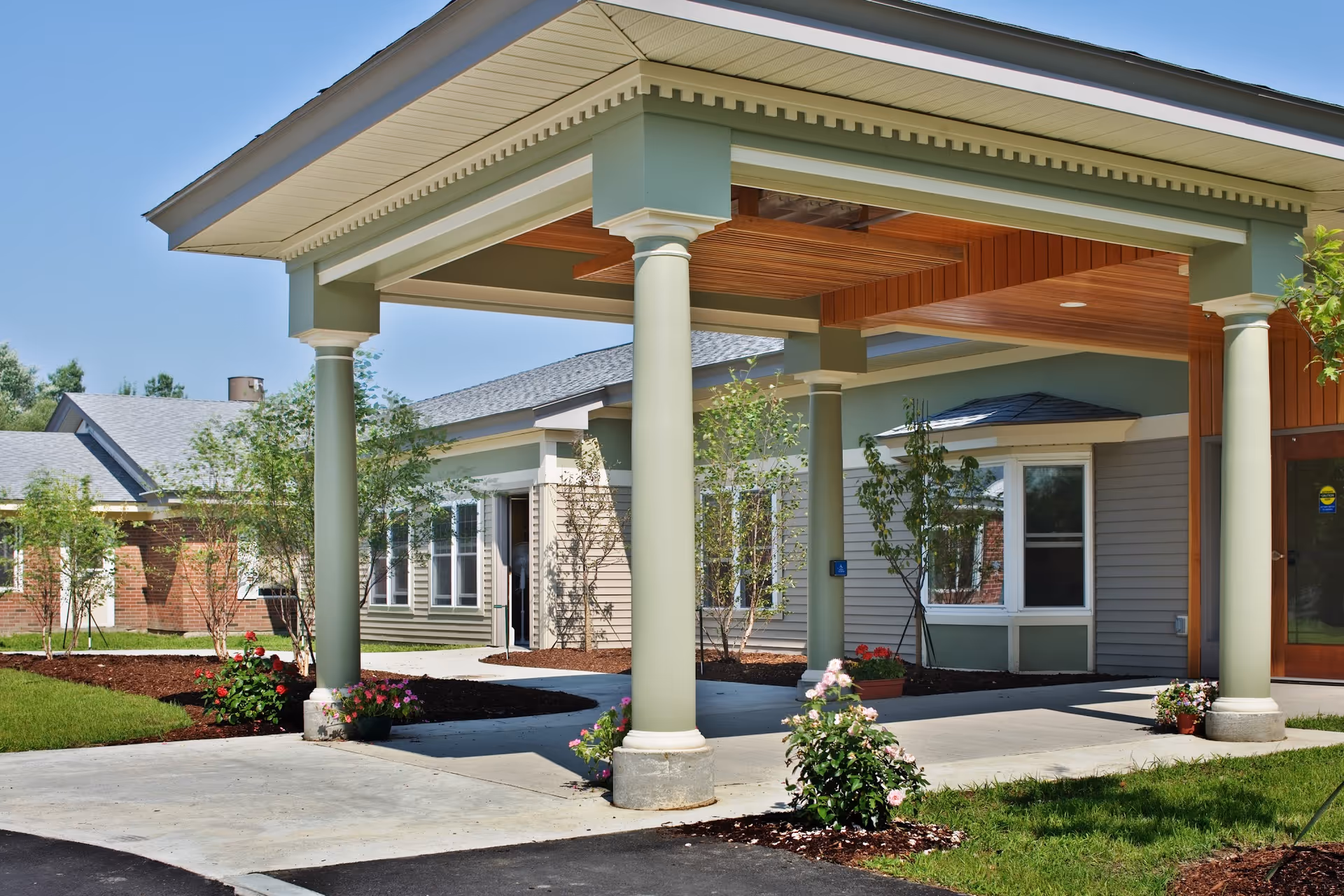 Covered porte-cochère with pale green columns, a walkway and landscaped flowerbeds at the front of a single-story residential building.