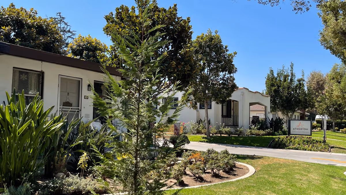 Landscaped front of the Atherton senior living facility showing white buildings, trees, lawn, and a sidewalk sign.